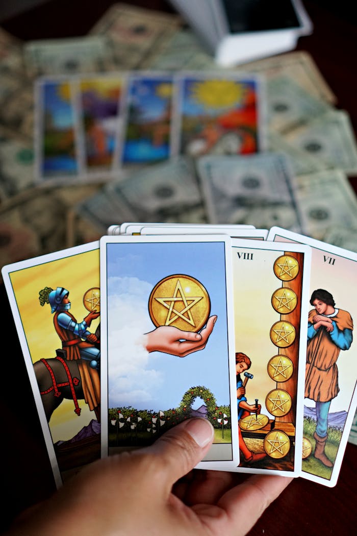 A hand holding tarot cards with money and tarot cards spread on a table.
